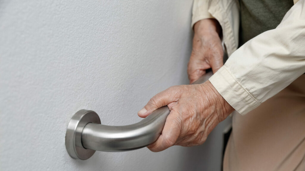 alt="a senior man's hands gripping onto a railing installed on a gray wall"